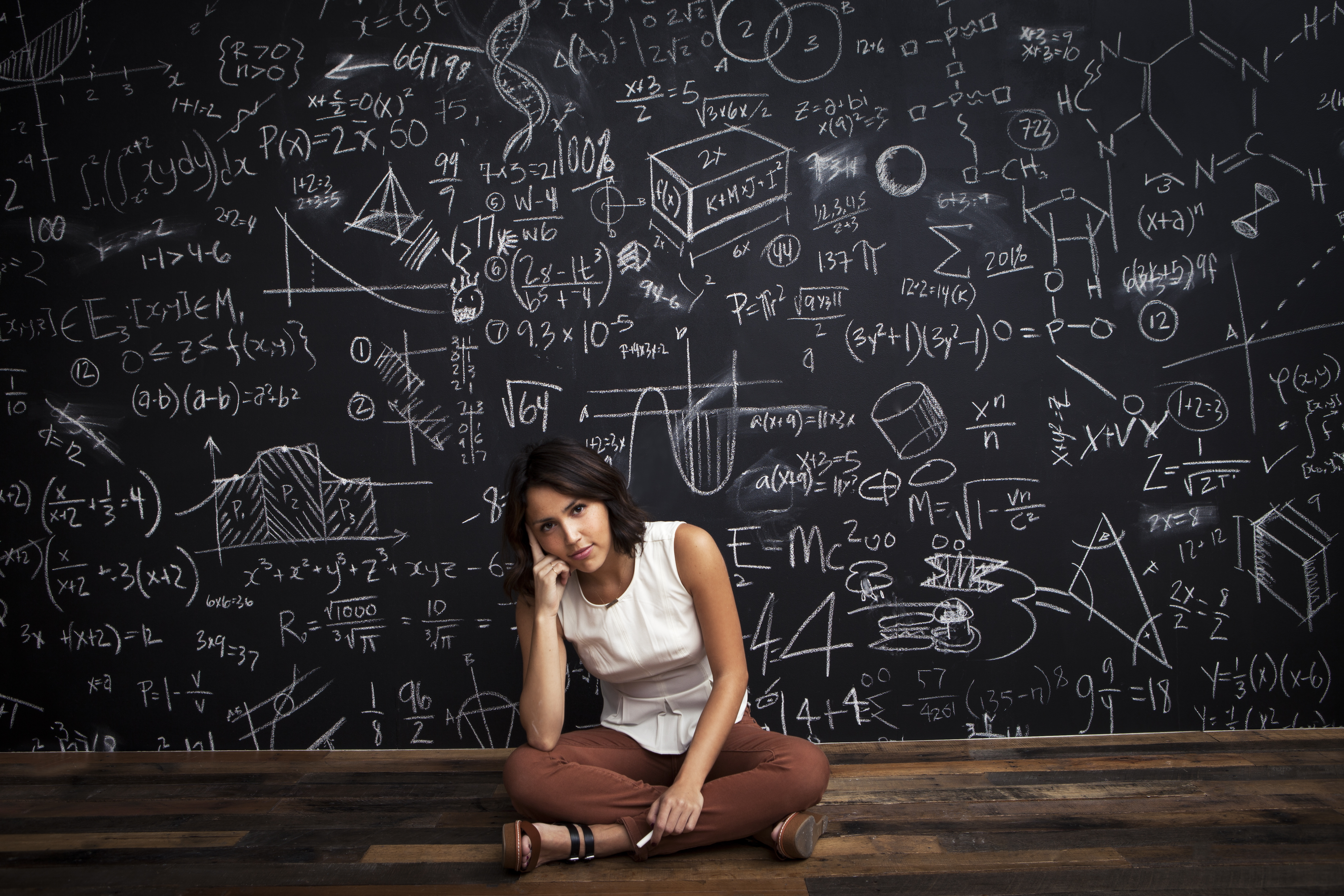 Woman sits cross-legged in front of a blackboard filled with mathematical equations and diagrams