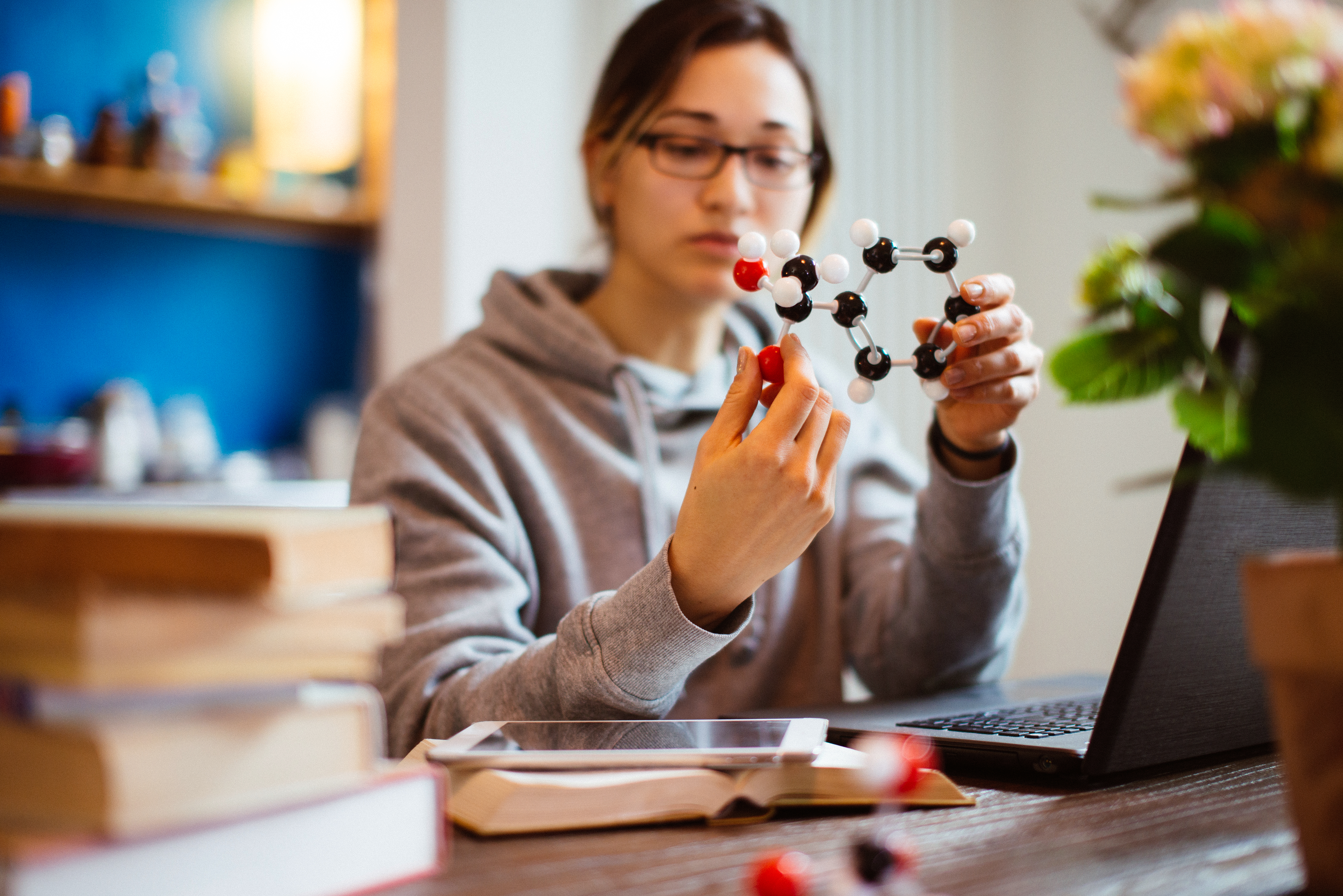 Woman examines a molecular model while studying at a computer with iPad and textbook close by