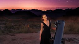 Raquel Nuno standing with a telescope in front of mountains and a starry sky Raquel Nuno standing with a telescope in front of mountains and a starry sky